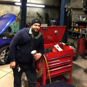 A burly mechanic in a blue hoodie leans on a red mechanics toolbox, ready to answer all your car maintenance questions!