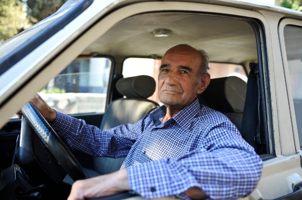 An elderly white man in a blue button down shirt sits in the driver's seat of a car.